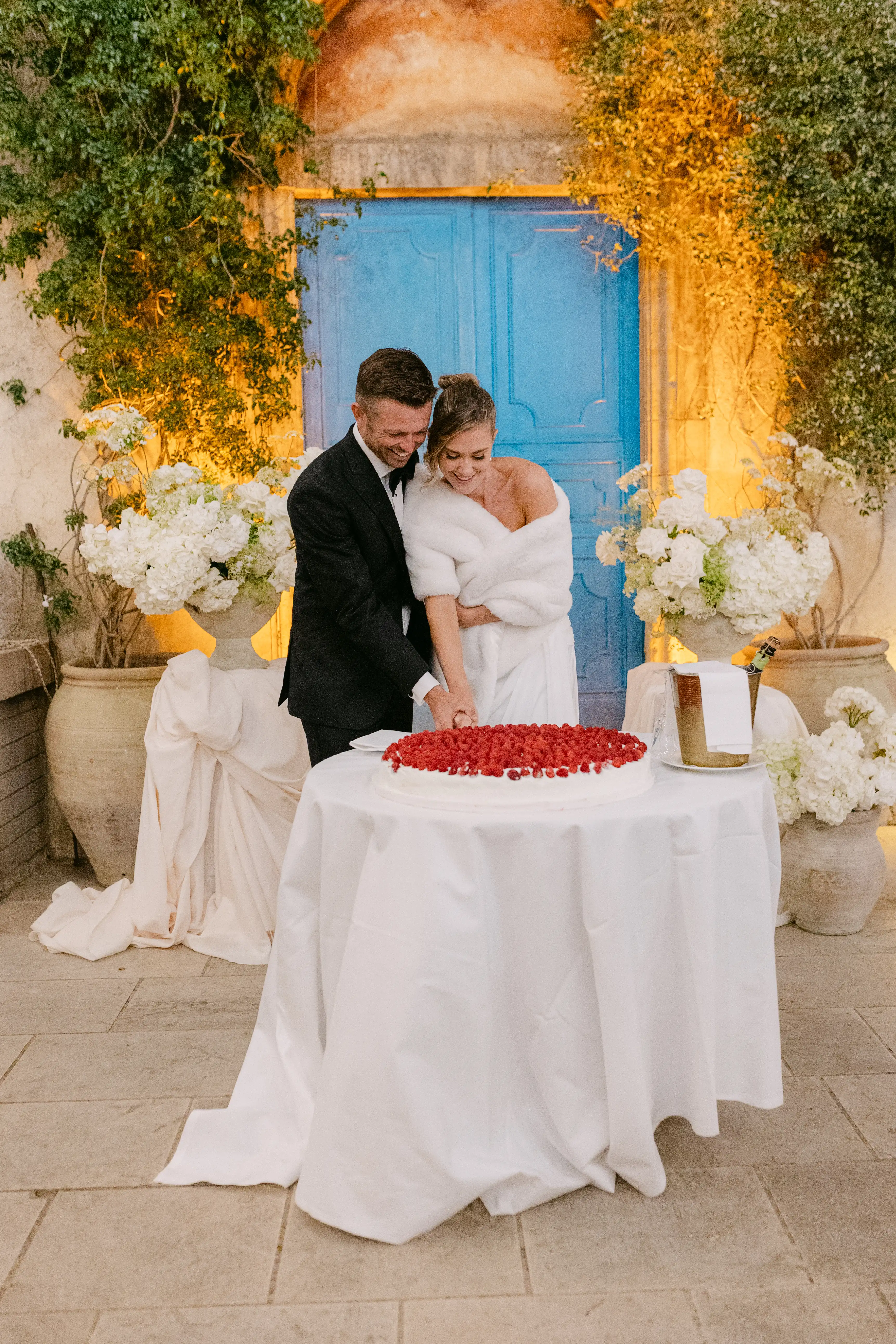 Bride and groom cutting a raspberry wedding cake in front of a blue historic doorway at a luxury Italian villa.